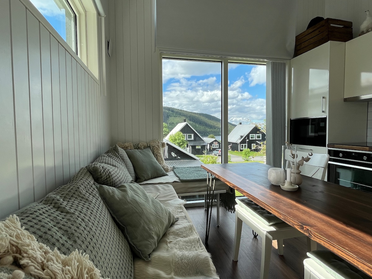 A bright dining area features a wooden table surrounded by white chairs. Natural light streams through large glass windows, providing scenic views of the surrounding hills. Textured cushions are arranged on a cozy sitting area along the wall, adding comfort to the space.