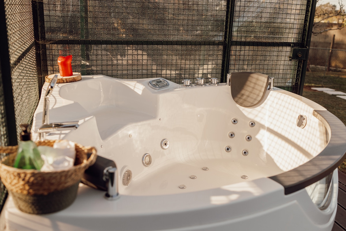 A modern jacuzzi sits in a spacious outdoor area, surrounded by a protective mesh fence. The inviting features include controls for water jets and a small basket holding toiletries. Natural light filters through, enhancing the relaxing atmosphere of the setting.