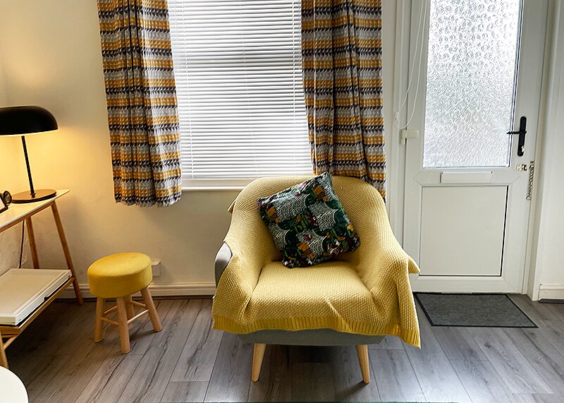 A cozy seating area is present, featuring a light grey armchair draped with a yellow throw blanket and adorned with a patterned cushion. Natural light filters through a window, complemented by patterned curtains. A small yellow stool and a side table sit nearby, enhancing the space.