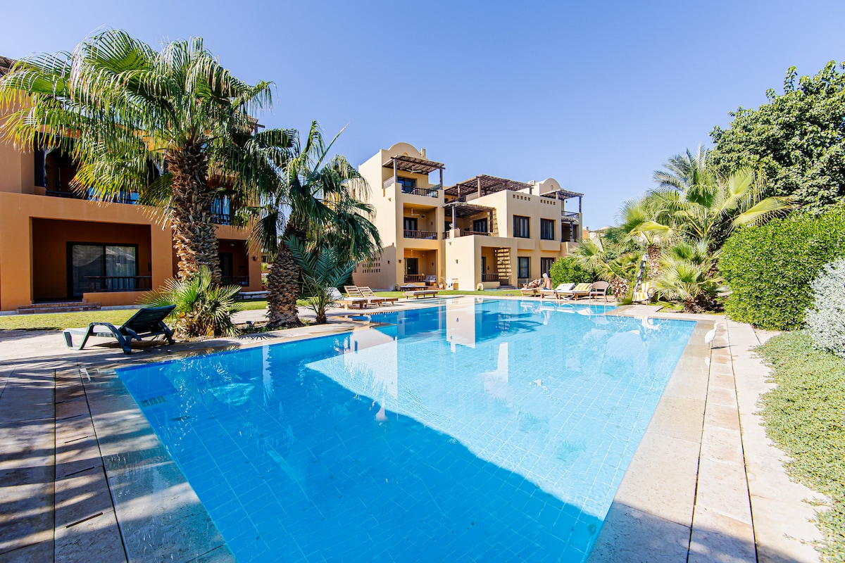A clear view of the communal swimming pool surrounded by lush palm trees and green foliage. Sun loungers are positioned around the pool's edge, inviting relaxation. The two-story apartment buildings in the background complement the tranquil outdoor environment.