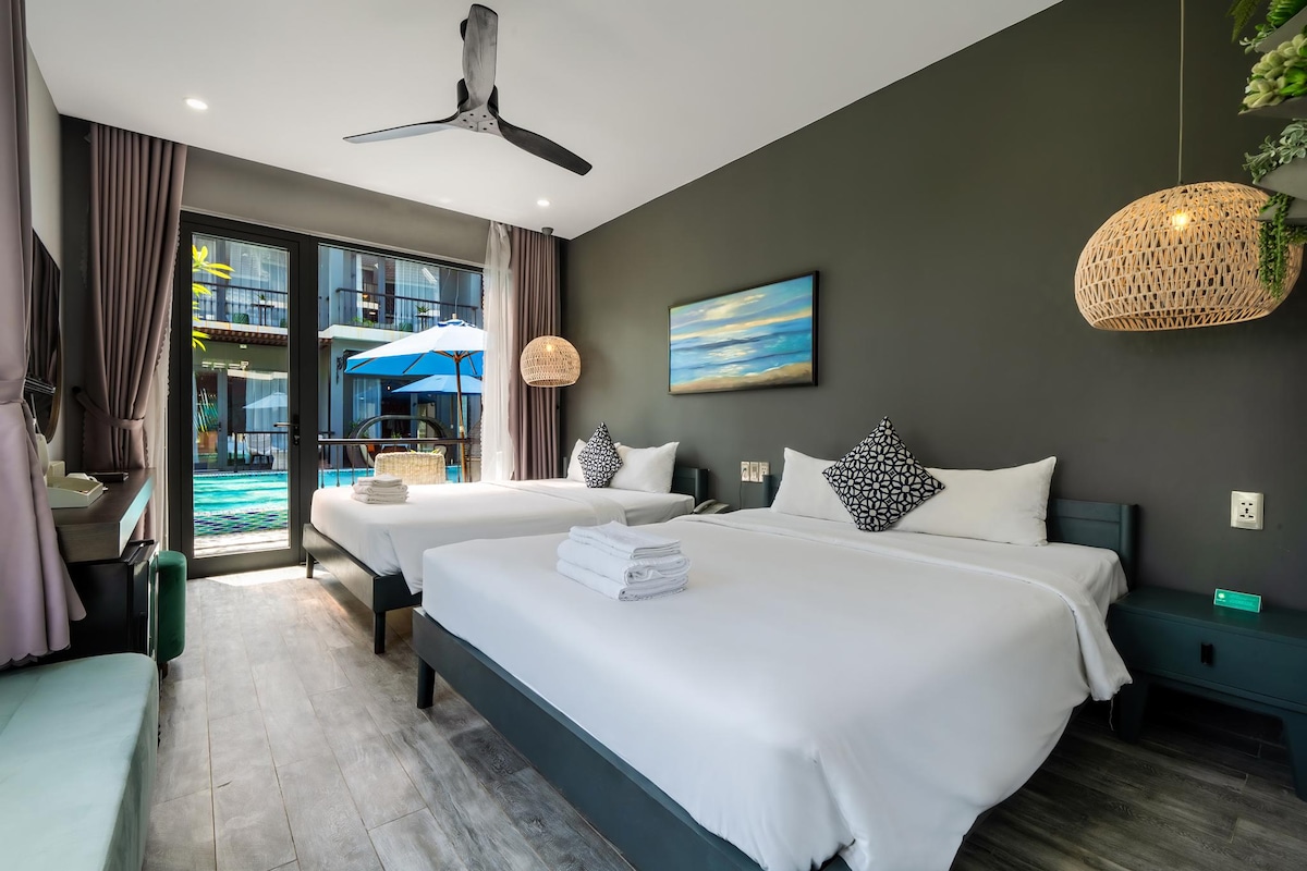 A bright and spacious guest room is showcased with two large beds dressed in white linens. Natural light fills the space through a glass door, which opens to a view of the pool area. Decorative wall art and light fixtures enhance the modern design.