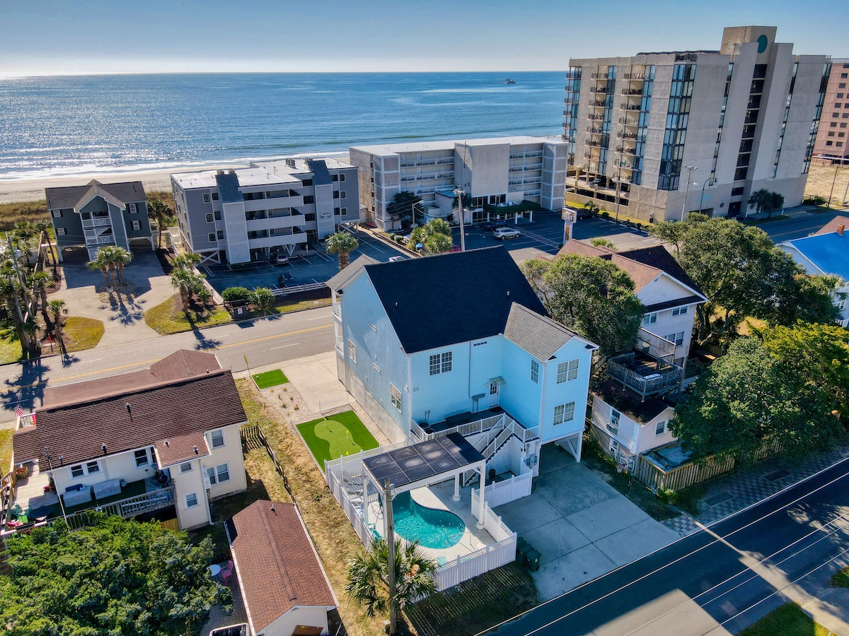 An aerial view captures the beach house nestled among surrounding properties, with the sparkling ocean visible in the background. The private pool is prominently featured in the backyard, bordered by a shaded seating area. The sandy beach is just a short distance away, promoting easy access.