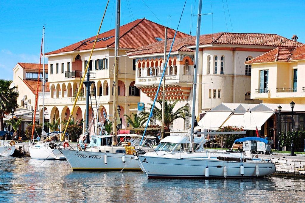 A marina setting features several sailboats docked in calm waters. Nearby, charming buildings exhibit a blend of architectural styles, complemented by palm trees and outdoor seating areas under canopies. Bright blue skies enhance the inviting atmosphere of this coastal locale.