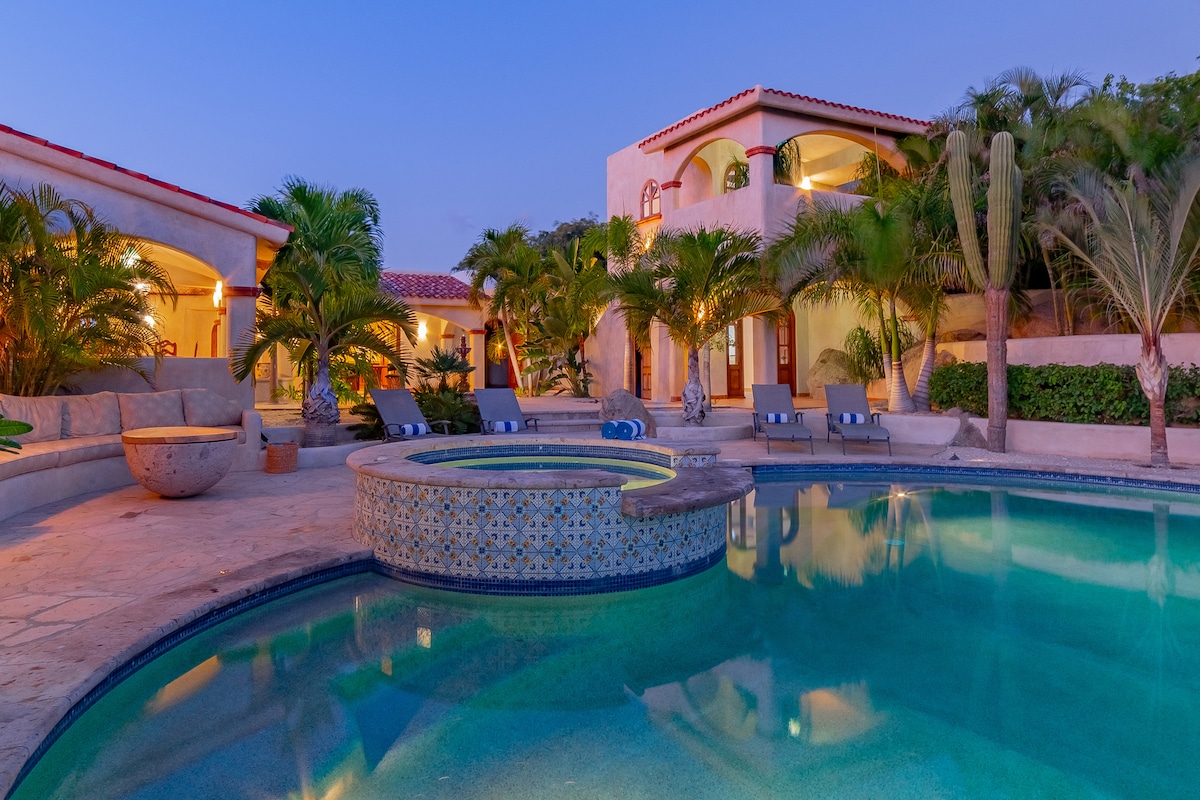 The outdoor area features a tranquil pool with an attached hot tub, surrounded by lush greenery and an elegant stone patio. The neighboring hacienda-style villa showcases arched doorways and scenic views, creating a serene ambiance as the sky transitions into evening.