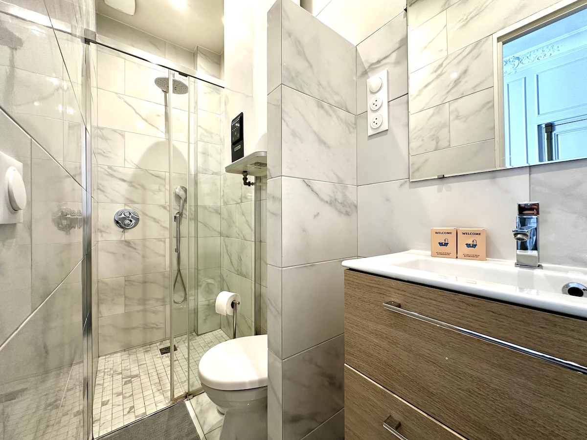 A modern bathroom features marble-patterned tiles throughout. A glass shower with a handheld showerhead is situated on one side, while a compact vanity with a sink and storage is positioned next to a toilet. Natural light illuminates the space through a small window.