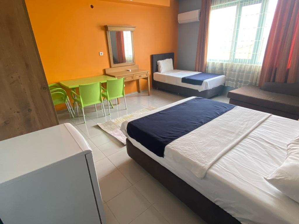 A bright room features an orange accent wall paired with a neutral color scheme. A spacious bed is positioned beside a single bed, with a comfortable sofa nearby. A small dining table with green chairs is present, alongside a compact refrigerator and a full-length mirror.