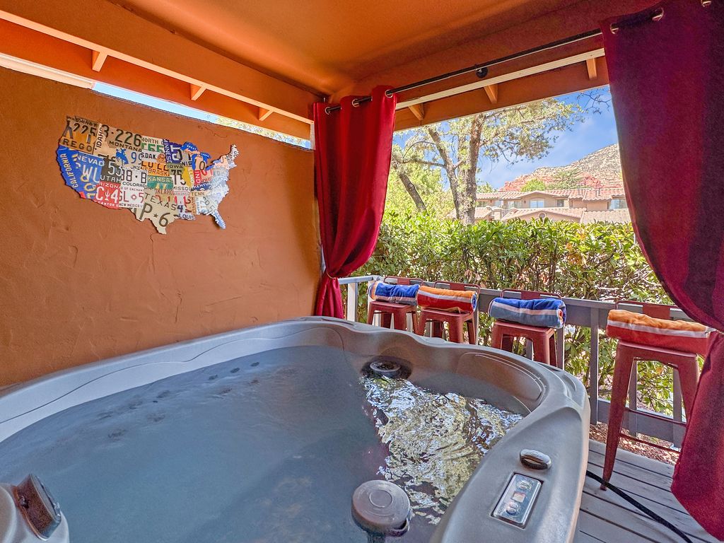 A private hot tub is featured on the front deck, with a view of the surrounding foliage and distant red rock formations. The area is adorned with vibrant curtains and seating that offers a comfortable spot to relax. A decorative map of the United States is displayed on the wall.