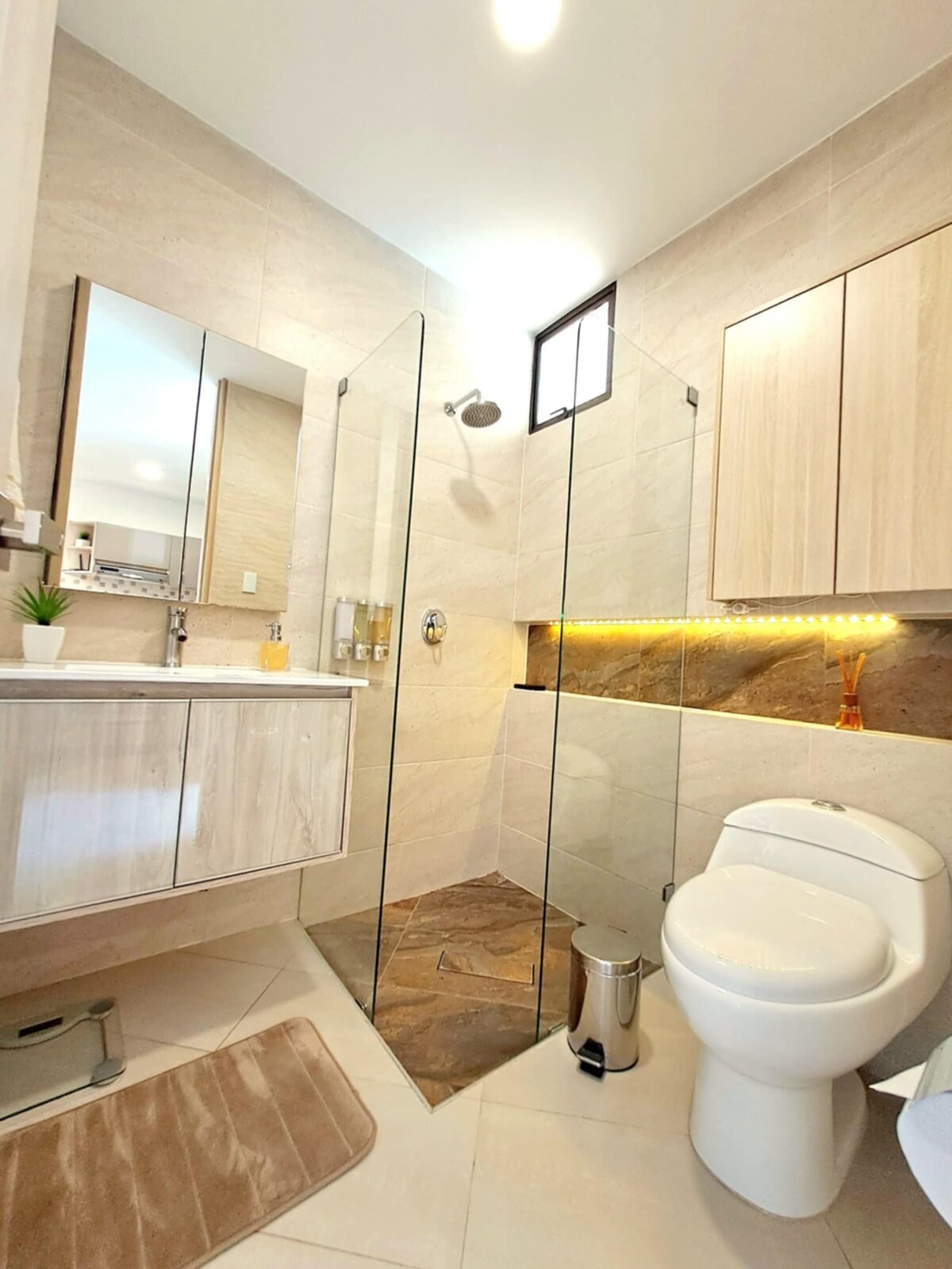 The bathroom features modern design elements, with a spacious glass shower enclosed by clear panels. A sleek vanity with ample storage and a mirror is positioned above a light-colored countertop. Warm lighting accents a tiled wall, enhancing the contemporary atmosphere.