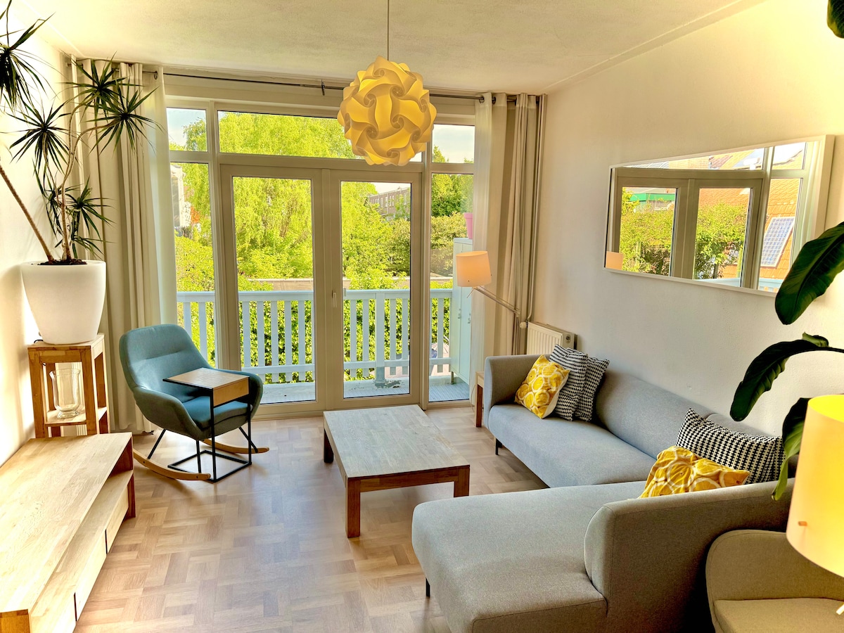 A bright living room features a spacious gray sofa and a wooden coffee table, complemented by a cozy armchair. Large glass doors provide access to a south-facing balcony, allowing natural light to illuminate the space. Green plants and decorative elements enhance the inviting atmosphere.