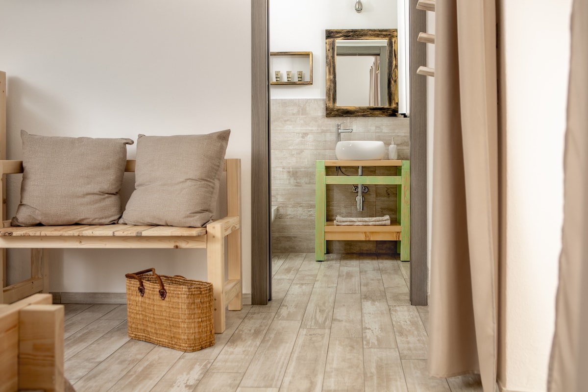A spacious room is shown with a wooden bench adorned with soft pillows. A light wooden floor complements the design. An open area leads to a stylish bathroom featuring a green sink stand and a large mirror above, creating a functional and serene space.