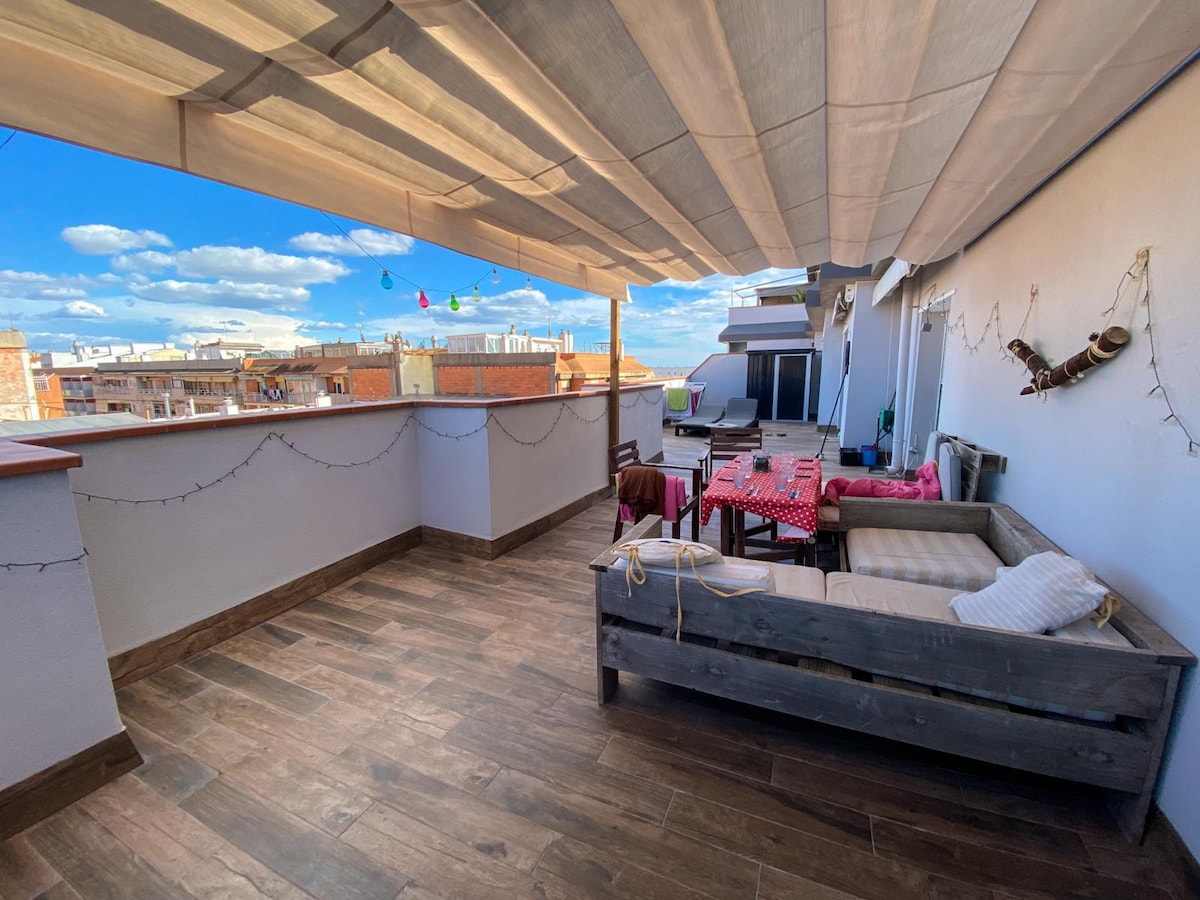 A spacious rooftop area is visible, featuring comfortable seating arrangements and a vibrant table set for dining. Decorative lights and colorful elements enhance the ambiance. The surrounding view includes rooftops and a clear sky, creating an open and airy feel.