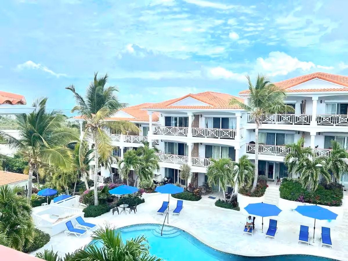 A serene view of the resort features palm trees and blue umbrellas surrounding a pool. The white building structure offers multiple balconies, and the lush greenery enhances the inviting outdoor space. Clear skies provide a bright backdrop to the tranquil scene.