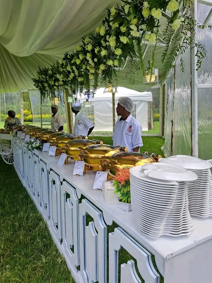 A buffet setup is displayed under a draped tent, featuring several golden serving dishes arranged along a long table. White plates are neatly stacked on one side. Decorative flowers hang from the tent overhead, contributing to a fresh and inviting environment.