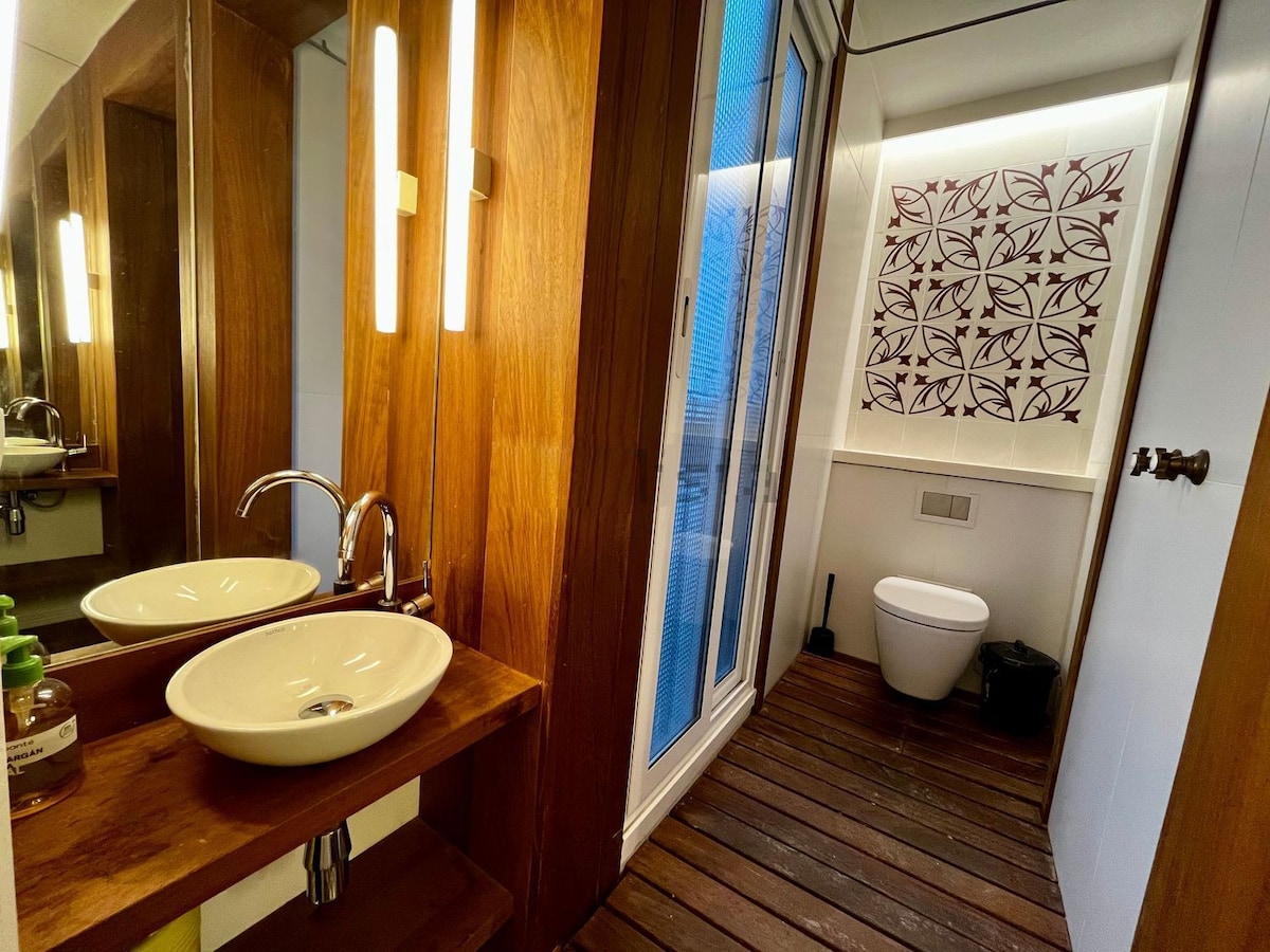 A clean and modern bathroom features two oval sinks on a wooden shelf. A large mirror is mounted on the wall, reflecting the warm wood tones. A toilet is positioned beside a door that leads to a small wooden balcony with a view.