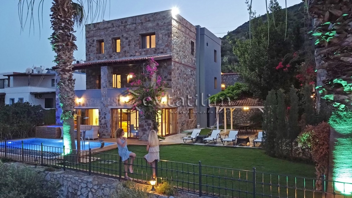 The exterior of Villa Torba Bay is highlighted by natural stone walls and vibrant landscaping. Two figures are seen near the illuminated pool area, while lounge chairs and a shaded terrace contribute to the outdoor amenities. Soft lighting enhances the overall ambiance as dusk approaches.