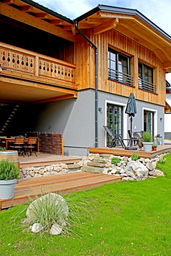 A two-story building showcases a blend of wooden and modern architectural elements. A spacious terrace is furnished with seating and an umbrella, while stone landscaping and green grass complement the exterior. A pathway leads to the entrance, enhancing the inviting ambiance.