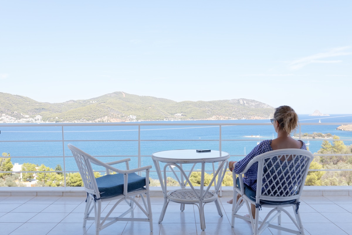A private balcony offers stunning views of the Aegean Sea and Poros Island. Two white wicker chairs with blue cushions and a round table create a serene space for relaxation, framed by expansive glass railings that enhance the unobstructed vistas of the surrounding landscape.