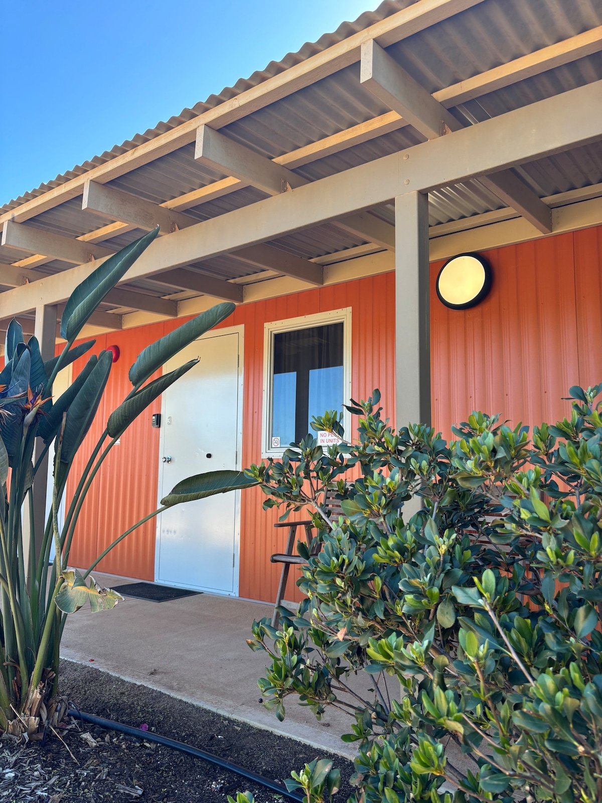 The exterior of a well-maintained accommodation features a vibrant orange facade complemented by a covered porch. Green foliage is present in the foreground, adding a natural touch. A secure entrance door and window are visible, providing direct access to the unit.