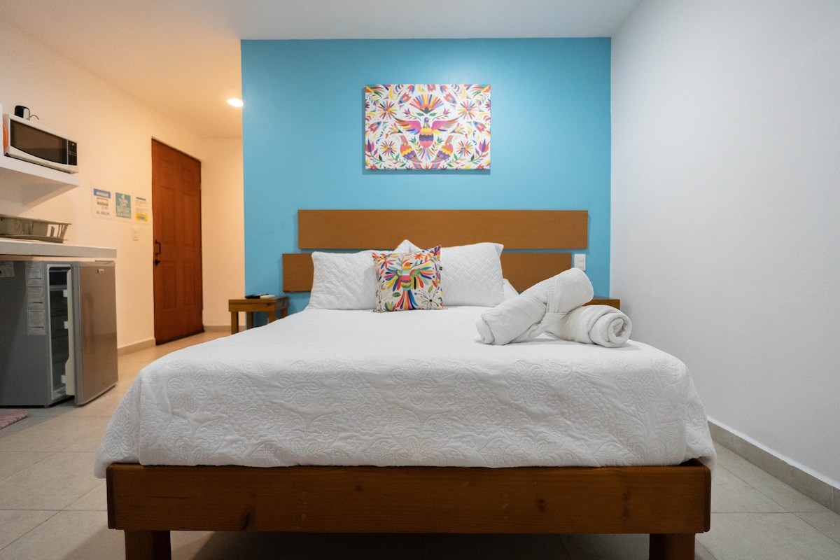 A cozy sleeping area features a spacious bed with a decorative pillow and neatly arranged towels. The wall behind the bed is painted in a calming blue, complemented by a colorful wall art piece. A small table and kitchenette are visible in the background.