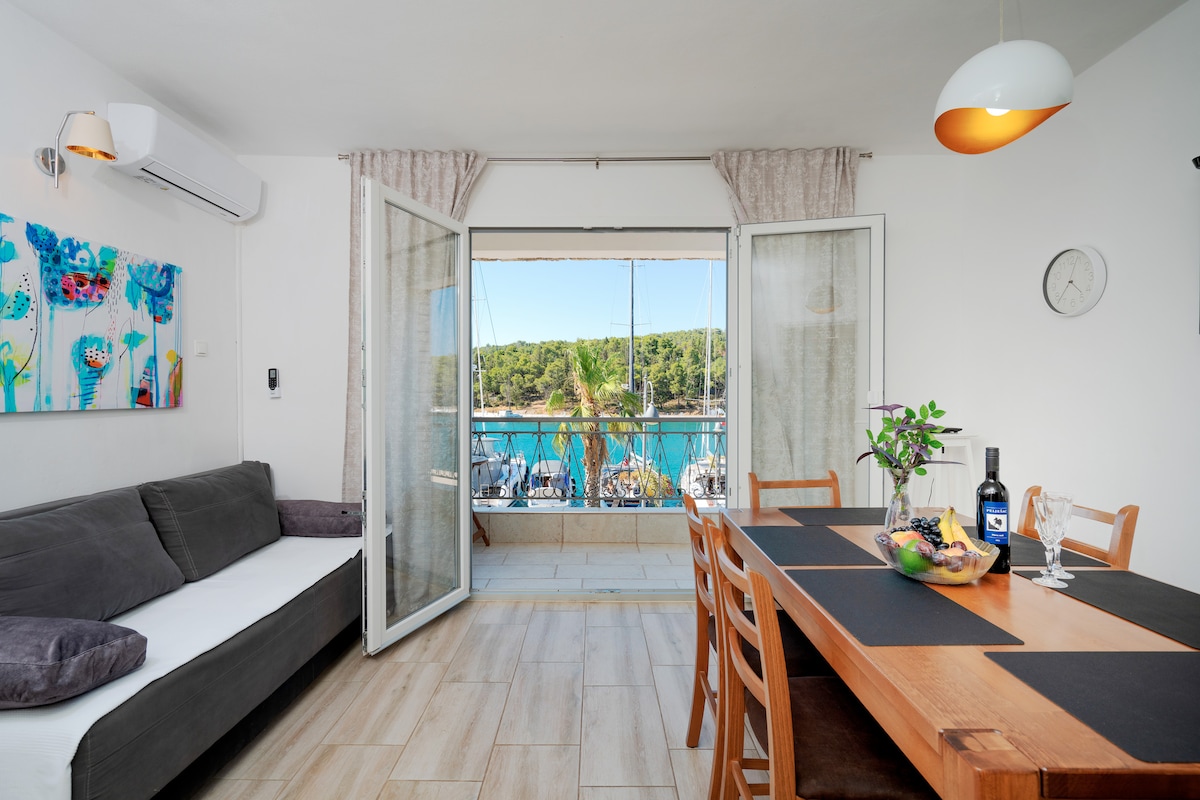 A spacious living area is presented with large glass doors that open to a view of the marina and lush greenery. A dining table is set for meals, complemented by a comfortable sofa adorned with cushions. Colorful artwork decorates one wall, adding a touch of vibrancy.