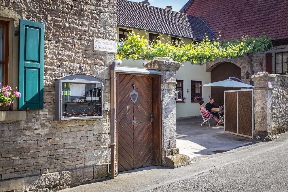 A welcoming entrance to a family winery is visible, featuring a rustic wooden door framed by stone walls. Potted flowers add color, while a courtyard area with seating invites guests to relax. Nearby, a modern building complements the historical architecture.