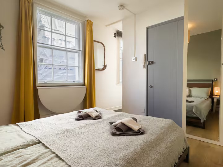 A cozy sleeping area is featured, showcasing a spacious bed covered with a light blanket. Two neatly folded towels are placed on the bed. Large windows with bright yellow curtains allow natural light to fill the space, while a mirror adds depth to the room.
