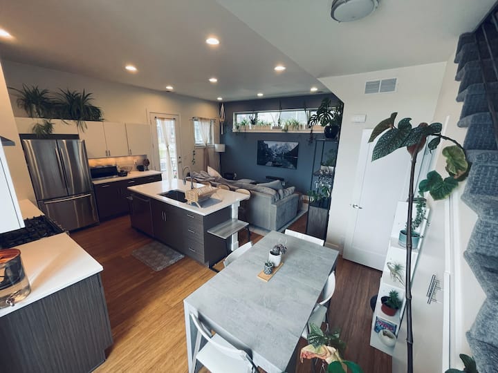 Кујна со отворен концепт, трпезарија и дневен простор со модерни завршни обработки и многу природна светлина и растенија. 