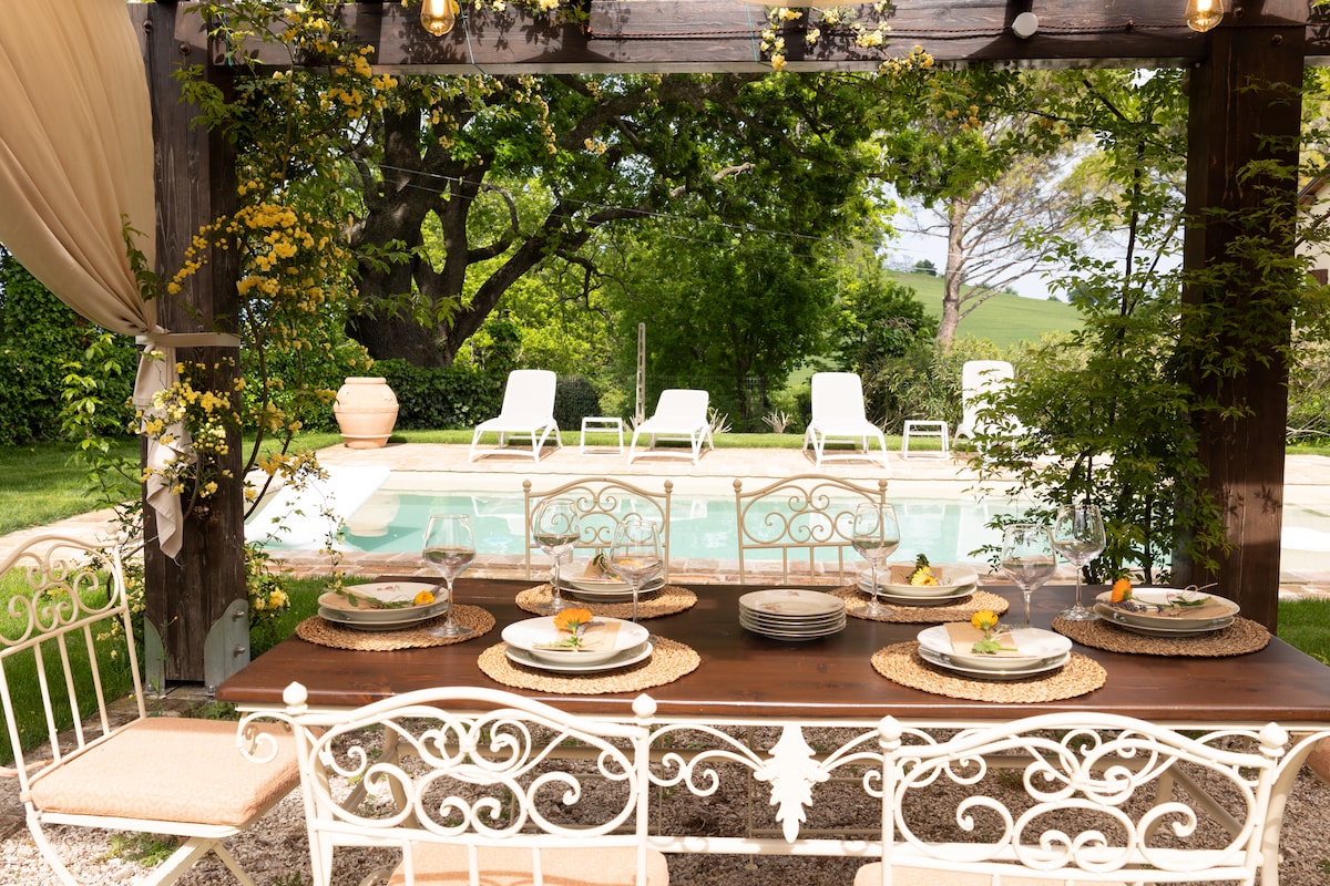An outdoor dining area features a long wooden table set with plates and glasses, surrounded by intricately designed white iron chairs. In the background, a sparkling pool is bordered by sun loungers, framed by lush greenery and soft sunlight filtering through trees.