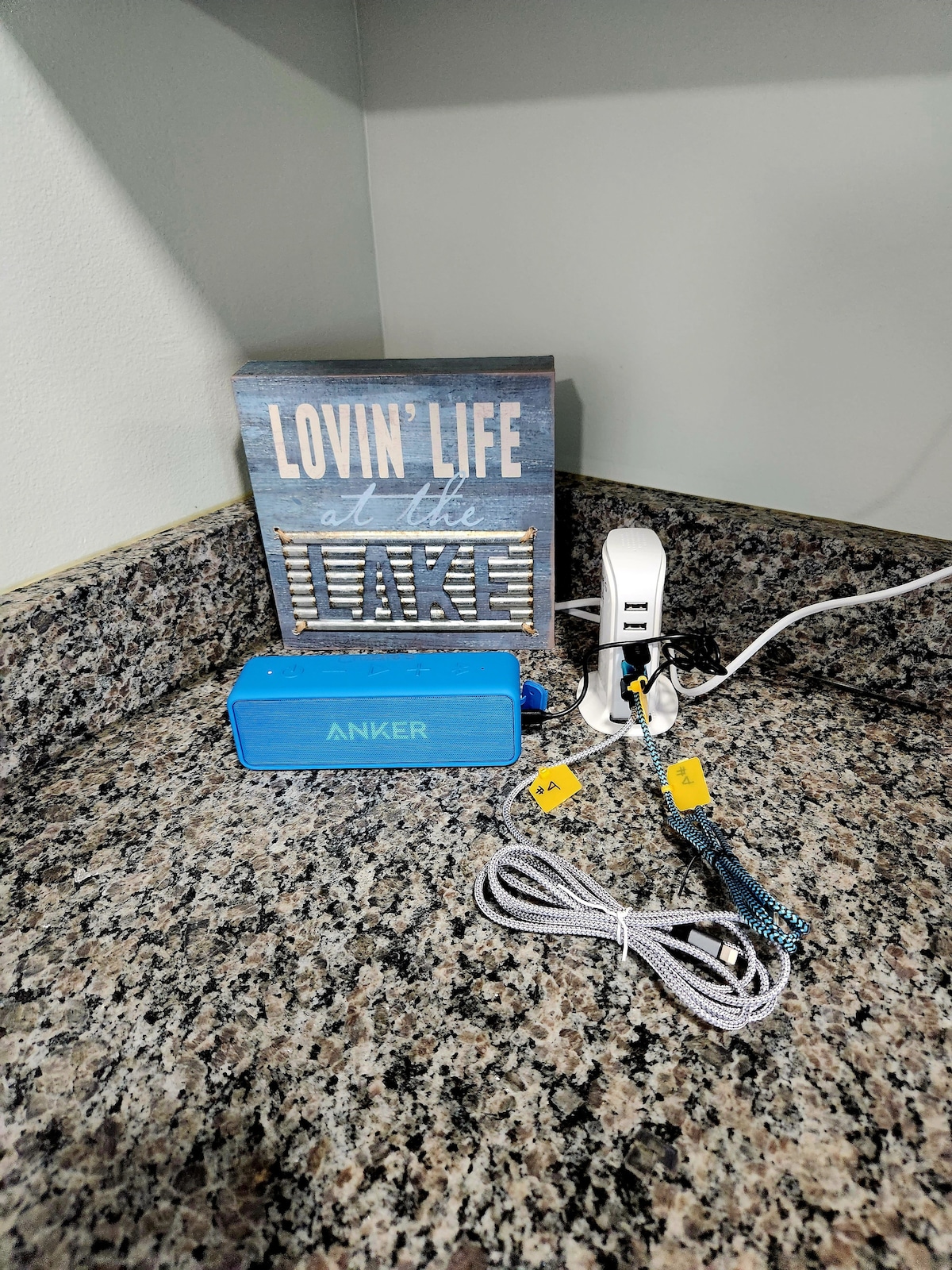 A countertop is displayed with a small Bluetooth speaker in blue, accompanied by a white charging station and power cords. A decorative sign featuring the words 'Lovin Life at the Lake' adds a cheerful touch to the organized space.