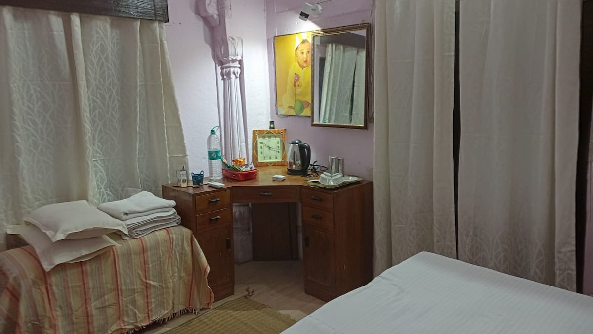 A cozy room features a wooden desk equipped with a kettle and various drinkware. Soft white curtains frame the window, complemented by neatly arranged towels on a bench. A decorative mirror above the desk enhances the room's ambiance, contributing to a space suitable for relaxation.