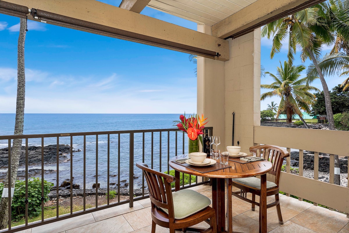A dining table set for two is positioned on a private lanai with a direct view of the ocean. Tropical plants and palm trees adorn the surrounding landscape. The table is complemented by two wooden chairs with green cushions, creating an inviting space for meals amidst nature.