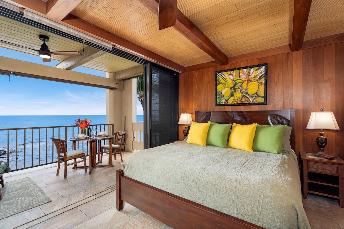 A spacious studio bedroom features a Cal king size pedestal bed adorned with a patterned green blanket. To the side, a small dining table with bamboo chairs is positioned near sliding glass doors that open to the ocean view. Teak-paneled walls add warmth to the space.