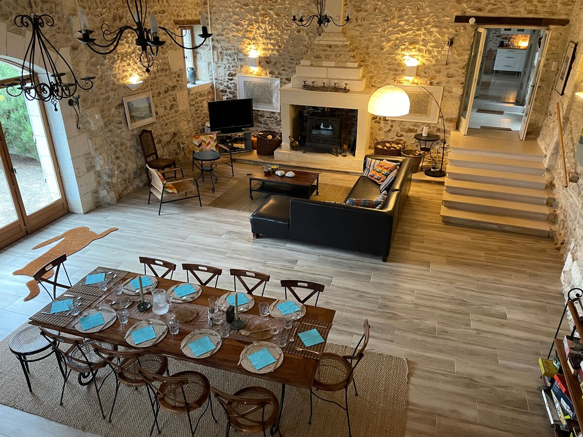 An open plan sitting and dining area features exposed stone walls and a wooden dining table set for a meal. A comfortable seating arrangement includes a large sofa and armchairs, with a television above the fireplace. Soft lighting enhances the welcoming ambiance.