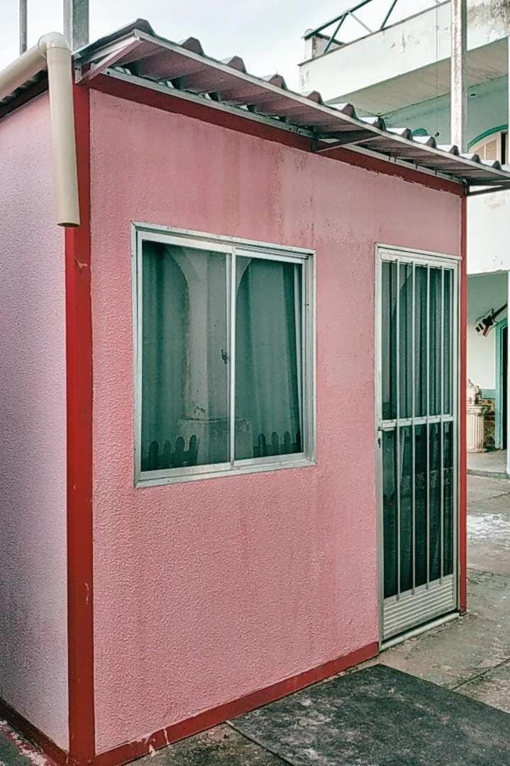 A small pink suite features a sliding glass door and a large window with a sheer curtain. The exterior is accented with red trim and a simple metal roof. Surrounding space is paved, providing easy access.