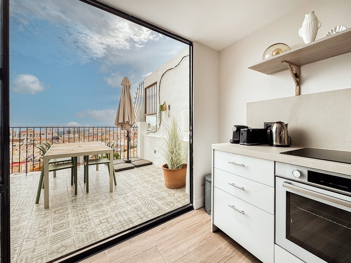 A modern kitchen space is viewed through expansive glass doors, which open to a terrace. The kitchen features sleek cabinetry and appliances, while a dining table with chairs is set on the terrace, complemented by a parasol for shade.