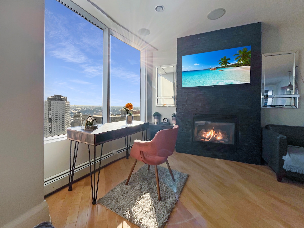 A bright workspace is positioned next to large windows, offering expansive views of the city skyline. A sleek desk is complemented by a pink chair, and a modern fireplace adds warmth to the space. A flat-screen TV mounted above the fireplace displays a serene beach scene.