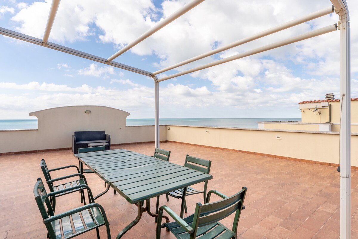 A spacious terrace offers a stunning sea view, featuring a large green table surrounded by six matching chairs. A comfortable seating area is positioned nearby against a wall. The blue sky is filled with soft clouds, creating a peaceful outdoor setting.