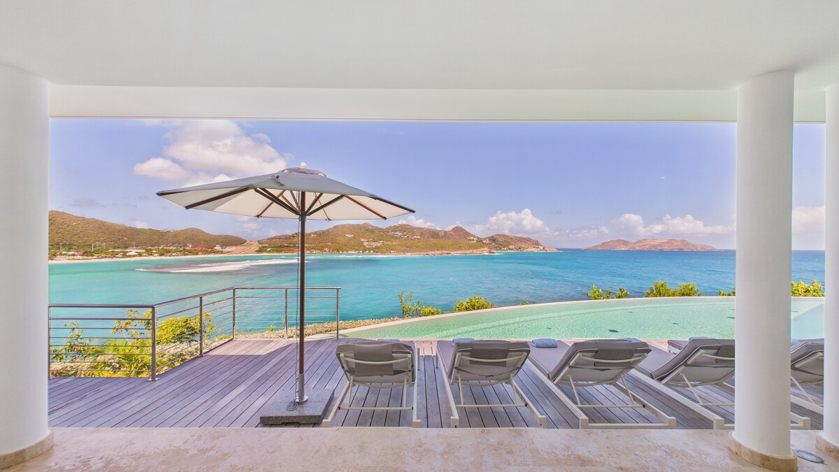 An outdoor terrace features lounge chairs positioned under a large umbrella, offering views of a vibrant blue sea and distant hills. The infinity pool reflects sunlight, enhancing the tranquil atmosphere. The white pillars of the villa frame this coastal scene, inviting relaxation.