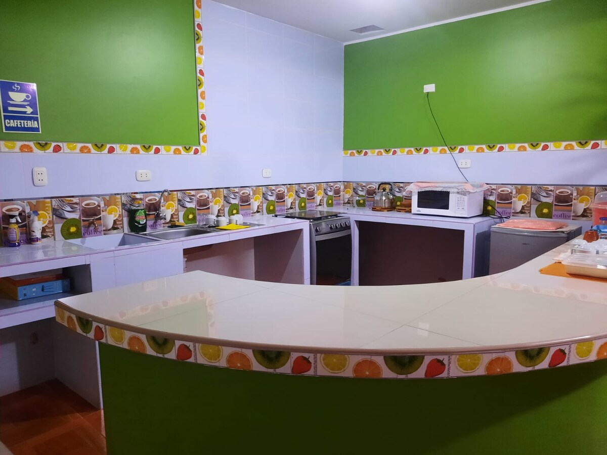 The kitchen features bright green walls and a smooth, white countertop that wraps around, offering ample workspace. Appliances include a microwave and a stove, with decorative tiles displaying fruits along the backsplash, contributing to a vibrant cooking environment.