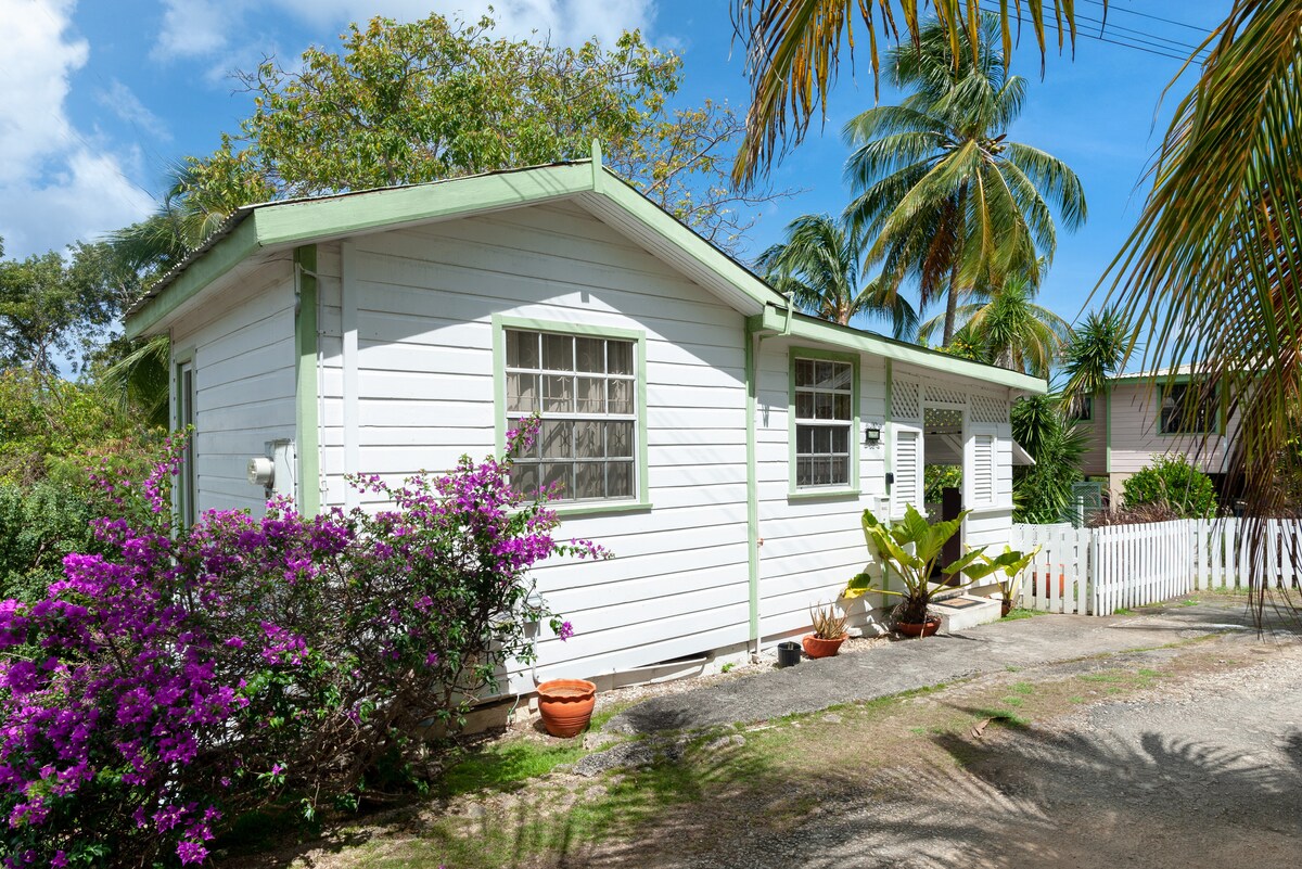 The exterior of the cottage features a charming white facade with green trim. Vibrant purple flowers are seen in the foreground, adding color to the scene. Palm trees surround the area, indicating a tropical climate, while a gravel path leads to the entrance.