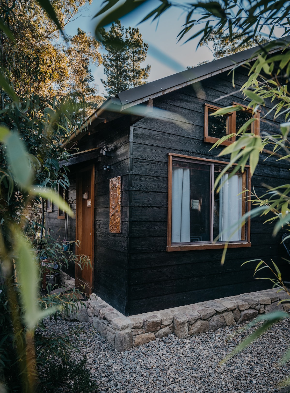 The exterior of the cottage is framed by lush greenery, showcasing a dark wood façade with unique window designs. A simple stone path leads to the entrance, where natural light filters through sheer white curtains.