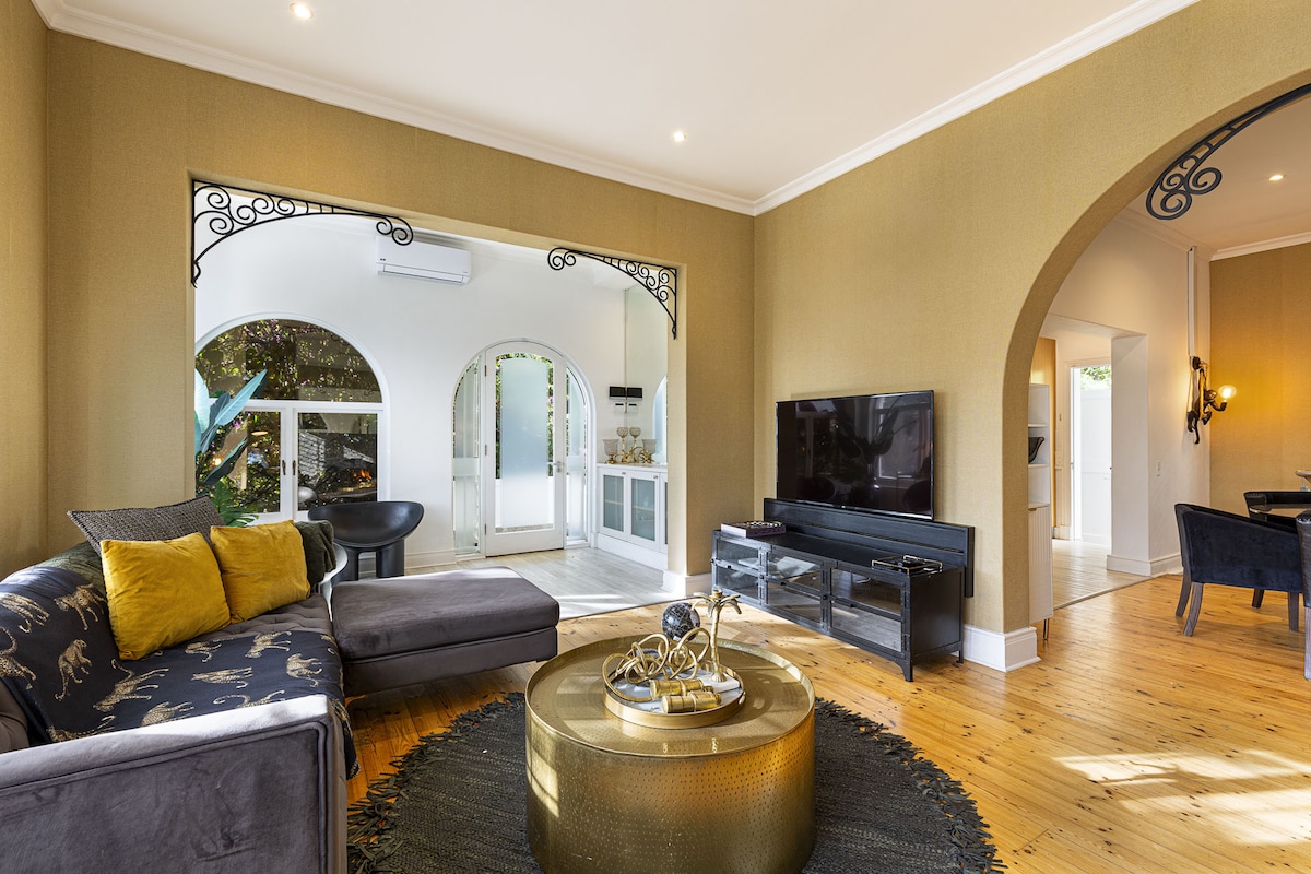 An open-plan living area features a plush sectional sofa adorned with decorative pillows, facing a flat-screen TV. Archways frame the space, which includes a round coffee table with a decorative centerpiece. Natural light fills the room, highlighting the wooden flooring and elegant decor.