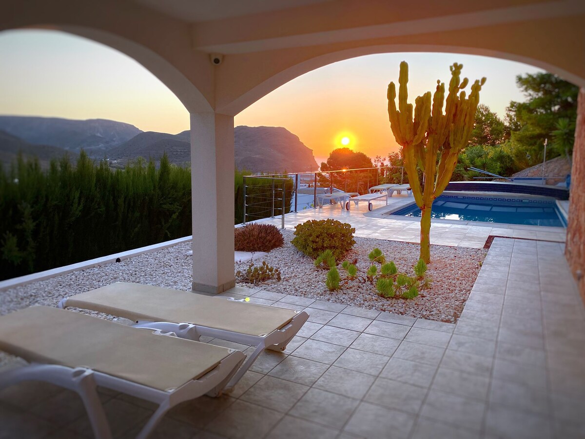 An outdoor area features a sun terrace with two loungers positioned on tiled flooring. Lush greenery and cacti adorn the space, while a shared pool is visible in the background. A sunset casts warm hues over the landscape, enhancing the serene atmosphere.