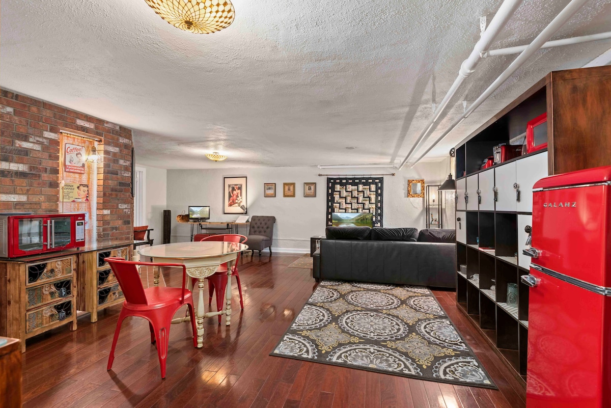A spacious living area is depicted, featuring a cozy sectional sofa, a circular dining table with red metal chairs, and a vintage red refrigerator. Textured wood floors and a patterned rug add warmth to the space, which is highlighted by exposed brick walls and thoughtful artwork.