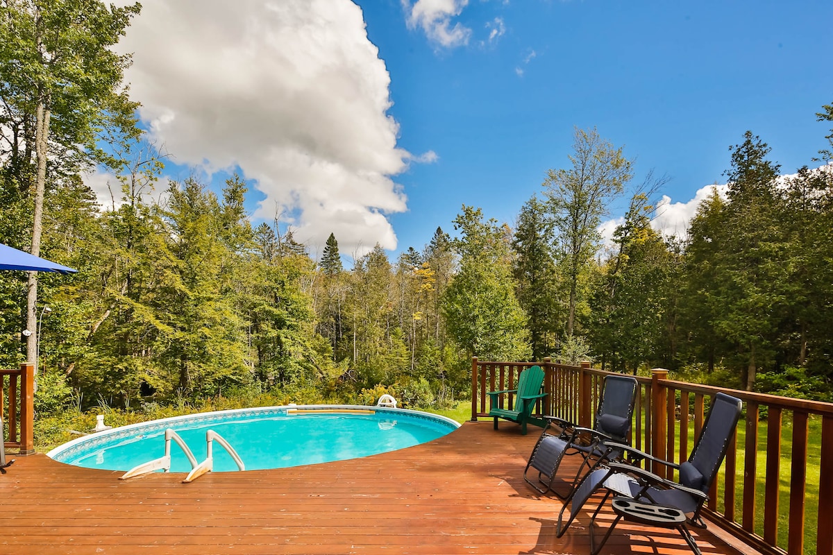 An outdoor deck surrounds a circular pool, providing access to the water via a set of steps. Two lounge chairs in dark colors and one in green are arranged on the wood surface, with a large umbrella casting shade nearby. Lush greenery envelops the area, creating a serene backdrop.