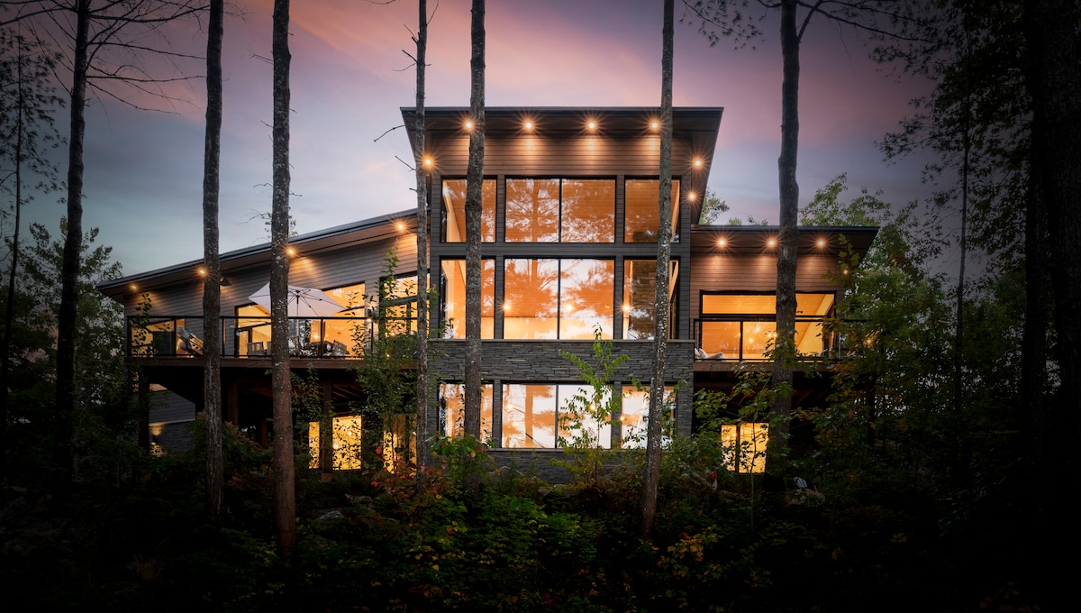 The exterior of the cottage is showcased amidst tall trees, featuring large windows that create a bright and airy appearance. Soft lighting highlights the architectural details, while the serene natural surroundings provide a tranquil setting.