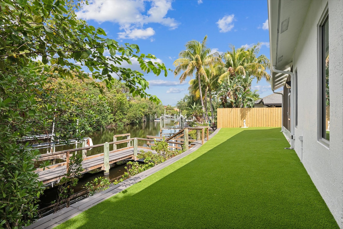 A lush outdoor space is presented, featuring a well-maintained green lawn that extends towards a calm waterway. Wooden decking is visible, offering access to the water, surrounded by dense tropical vegetation and palm trees under a clear blue sky.