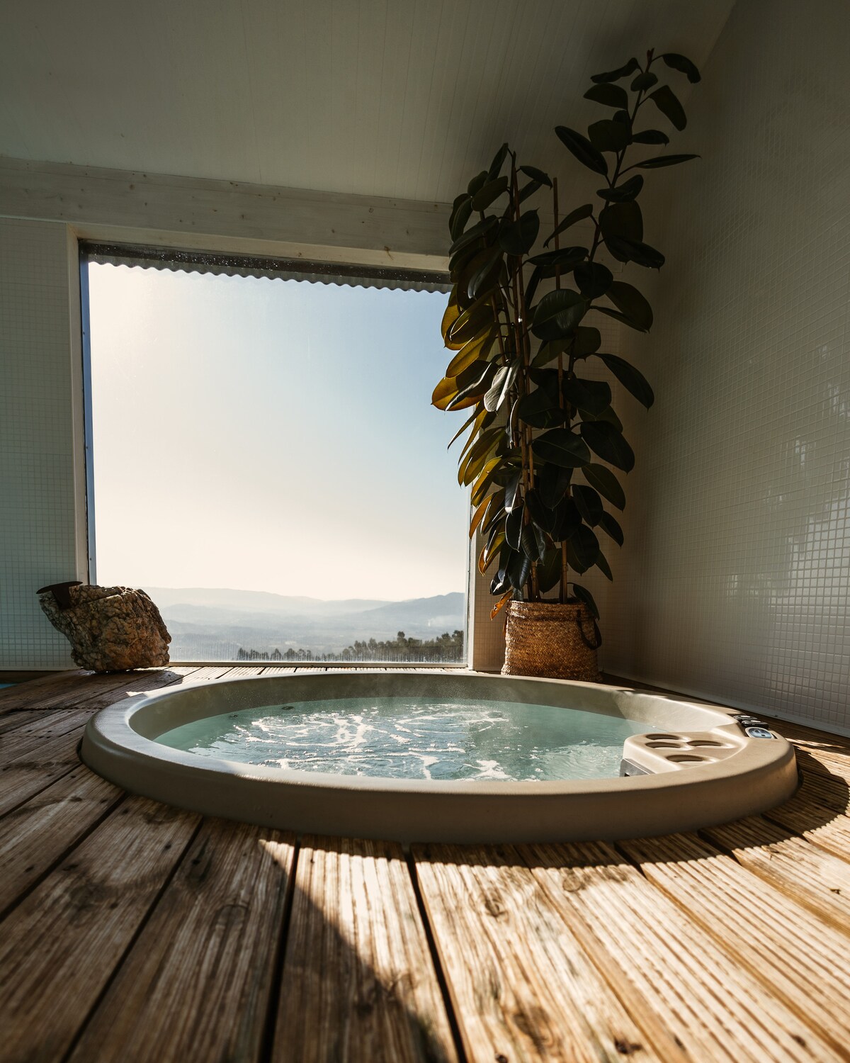 A jacuzzi is positioned by a large window, allowing for panoramic views of the mountainous landscape. Sunlight softly illuminates the space, highlighting the wooden flooring and a potted plant beside the jacuzzi. The atmosphere is serene and tranquil.