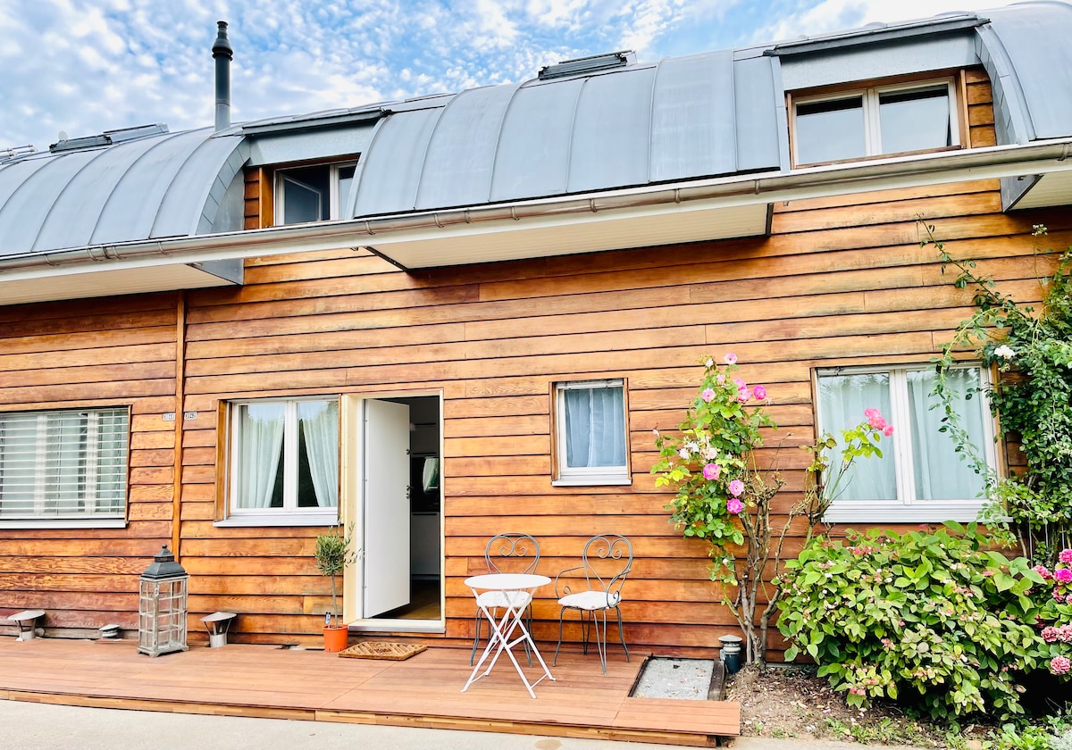 The exterior of the building features a warm wooden facade with a distinct metal roof. A small wooden deck includes a bistro table and two chairs, surrounded by greenery and pink flowers, creating a charming entryway. Windows are dressed with light curtains, allowing natural light to brighten the space.