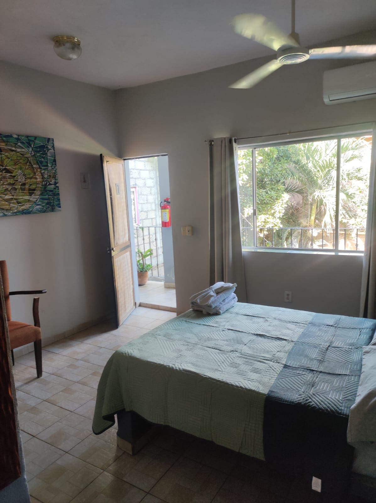 A bright room features a comfortable bed with a light blue blanket, complemented by a well-placed chair. Natural light enters through a window overlooking a lush outdoor area, creating a serene atmosphere. A door leads to a small balcony with potted plants.